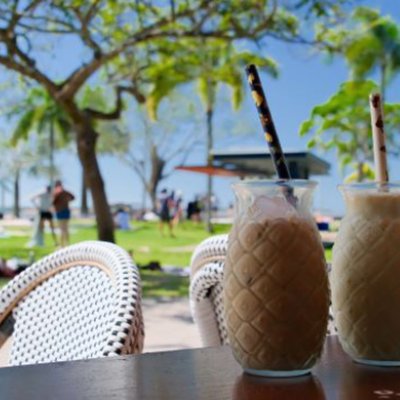 Two milky drinks with straws sit on a table in front of a green park with many people